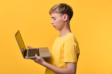 Young boy in a yellow shirt holds an open laptop against a bright yellow background, exploring digital learning and technology, with a focused expression and a modern, minimal setup that suggests