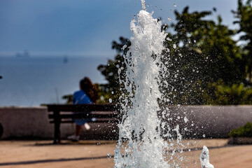 Obraz premium Close-up of a sparkling fountain jet with water droplets, offering a sense of cool refreshment on a hot day, with a person on a seaside bench in the background. Antalya, Turkey, Mediterranean.