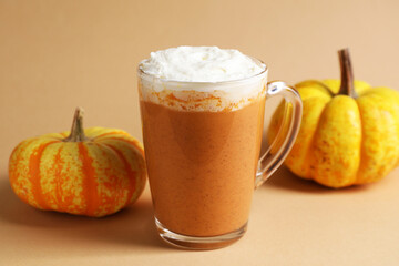 Delicious pumpkin latte with whipped cream in glass cup and fresh vegetables on beige background, closeup