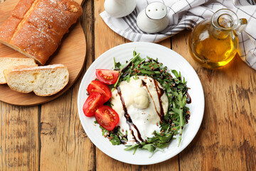 Delicious burrata cheese with arugula, tomatoes and dressing on wooden table, flat lay