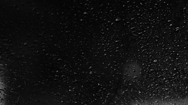 Extreme close-up cinematic black and white view of raindrops on a car window at night. Moody, abstract and atmospheric textural footage with high contrast detail