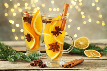 Delicious mulled wine with slices of orange, spices and fir branches on wooden table against grey background with blurred Christmas lights, closeup. Bokeh effect