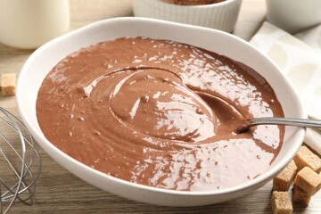 Liquid chocolate dough in bowl, whisk and brown sugar on wooden table, closeup
