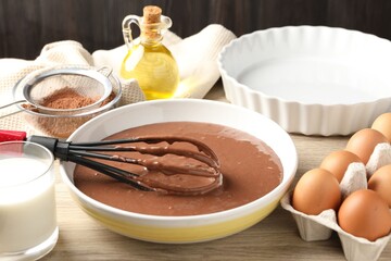 Liquid chocolate dough in bowl, whisk, oil, milk, cocoa powder and eggs on wooden table, closeup