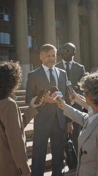 Vertical shot with lens flare of professional Caucasian male barrister with bodyguard getting interviewed by reporters holding microphones and recorders by court hall after lawsuit hearing