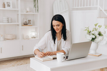 Young brunette freelancer woman sitting at desk using laptop writing notes while studying online, watching webinar, looking at pc screen learning web classes or remote working from home. Side view