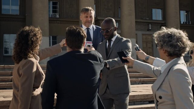 Full shot of middle-aged Caucasian male politician, dressed in blue business suit, walking through crowd of reporters after court case, black male security guard pushing through and protecting client - Powered by Adobe
