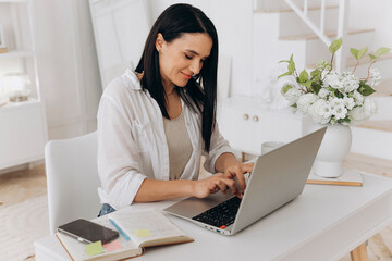 Freelance woman smiling while typing on laptop during virtual consultation, neat planner and phone nearby, airy home setting with decorative flowers, professional warm tone conveying client trust