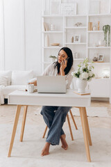 Caucasian freelancer woman on phone, remote project manager taking client notes at bright white desk with laptop, mug, minimalist bookshelf and natural light, focused professional mood
