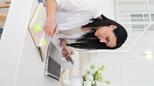 Intent caucasian woman arranging sticky notes and checking phone beside laptop, hands marking planner on bright desk, focused project manager vibe in minimalist room, organized task planning scene