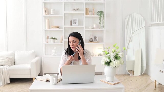 Consultant caucasian woman on phone at desk with laptop and notebook, animated hand gestures show negotiation and multitasking in bright living workspace, professional remote consultation vibe