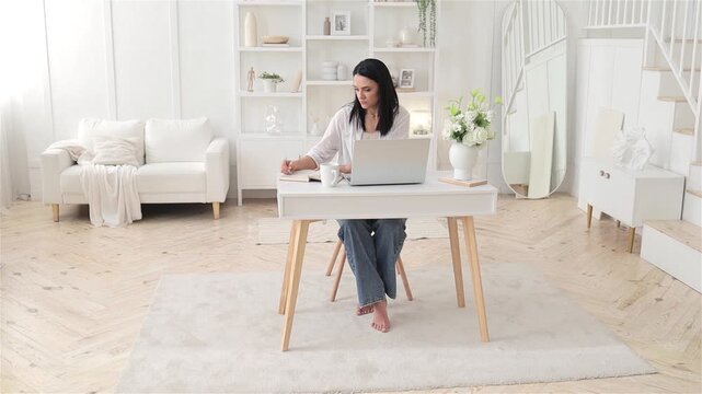Remote worker caucasian woman composing notes at desk with laptop and planner in bright minimal room, steady focused posture and calm atmosphere convey disciplined daily routine and productivity