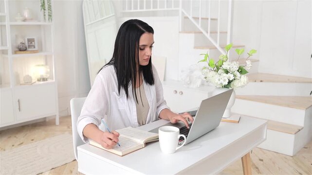 Content creator caucasian woman writing in notebook beside open laptop and mug, bright airy staircase backdrop with flowers, thoughtful research and planning for blog or project, quiet focused mood