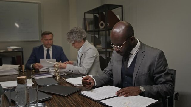 Medium shot of young African American male lawyer sitting at table in conference room during team meeting, analyzing and signing document drafts, while colleagues are discussing evidence files