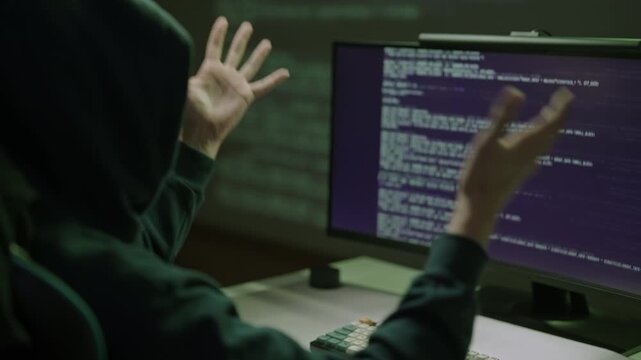 Anonymous person wearing hoodie raising hands in frustration while looking at malfunctioning code on computer monitor, indicating issues with programming, cybersecurity or data breach attempts.