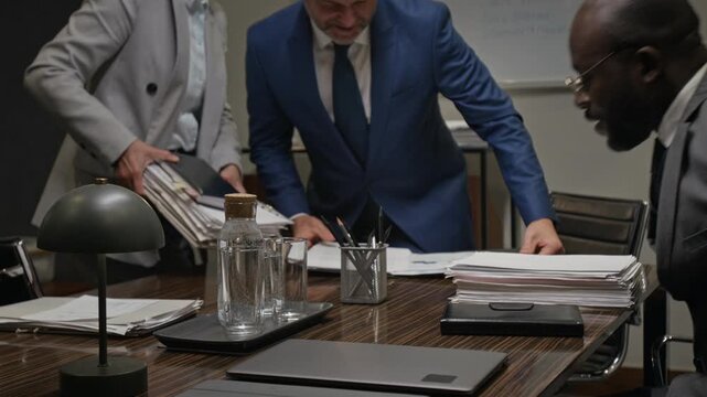 Medium shot of three diverse male and female law firm partners, dressed in business suits, entering conference room, sitting down for team meeting, opening notebooks and beginning case discussion