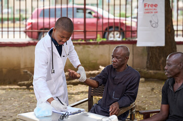 A young African male doctor in a white coat checks an older man’s wrist and writes notes while another man sits beside them during an outdoor free medical checkup.