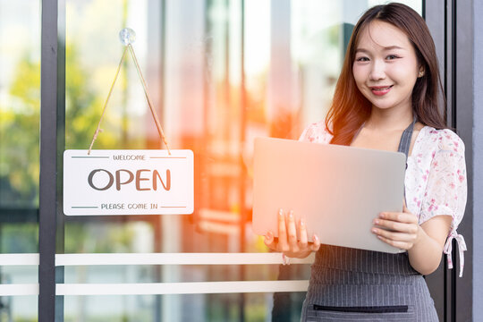 Female owner of coffee shop or restaurant turning round sign to open. Smiling young asian woman owner, employee retail coffee shop woman hand in setting sign board to open for welcome customer, reopen - Powered by Adobe