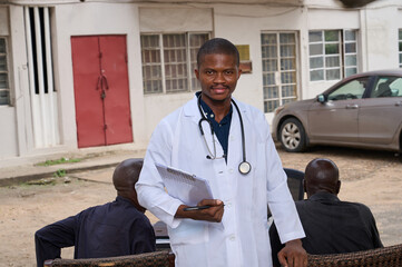 A male African health worker in a white coat stands outdoors holding a clipboard and pen, facing...