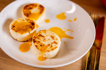 Description (≤200 chars):
Close-up view of cottage cheese pancakes with golden crust and bright pumpkin sauce on a white plate, styled with warm tones and gold cutlery.