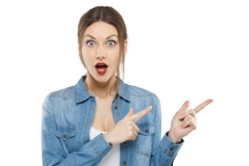 Portrait of a shocked young woman in a denim shirt pointing with both hands to the side