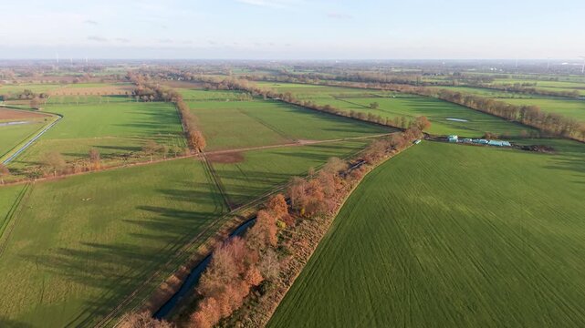 Aerial view of rural fields with crop rows, tree shadows, canal, and farm buildings sunlit landscape blending agriculture, infrastructure, and natural geometry.
