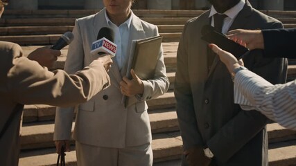 Medium shot of mature Caucasian female lawyer, wearing grey trouser suit, with documents folder, accompanied by bodyguard, refusing to speak to mass media journalists after court case hearing