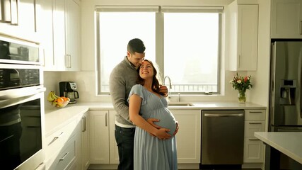 Joyful expectant parents cuddling and smiling in their bright and airy modern kitchen at home