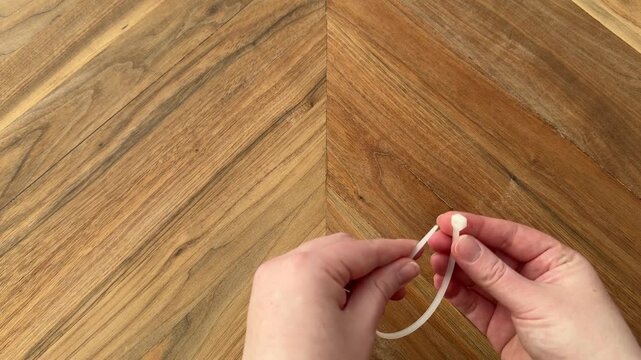 Female hand picking up a plastic zip tie, fastening it closed, and placing it down on a wooden table. 4K video.