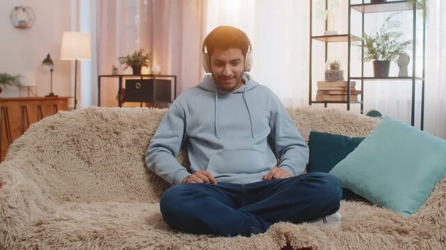 Indian young man sitting on sofa couch listening to music through headphones dances to rhythm relaxes and unwinds. Hispanic guy enjoys beats moving with energy and finding joy in favorite tracks.