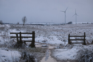 Winter rural landscape with a snow covered path, wooden gates and wind turbines in the distance. Cold countryside scene showing renewable energy and seasonal weather.