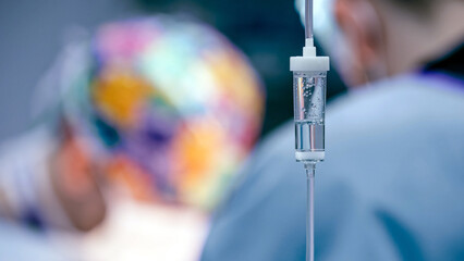 Medical IV drip stands prominently in the foreground, with blurred medical professionals in...