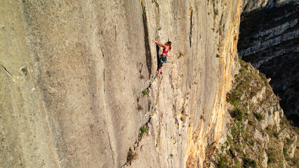 AERIAL: Young woman climbs a tall limestone wall high above the Cikola canyon in Croatia. Scenic view reveals the sheer cliff, tiny climber and dramatic exposure of this natural rock climbing area.