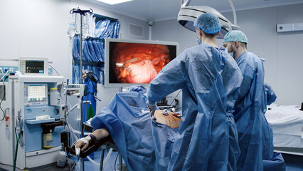 Medical professionals in surgical attire are performing a complex operation in a modern operating room, with advanced equipment and a large monitor displaying vital information
