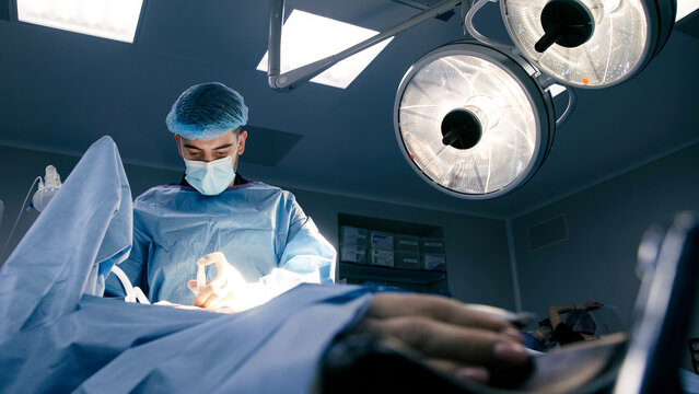 Male surgeon in blue scrubs and surgical mask is performing an operation under bright surgical lights, showcasing precision and focus in a modern operating room environment