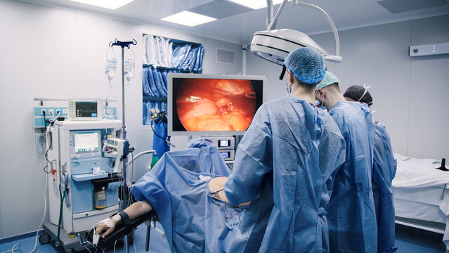 Medical team of surgeons in blue scrubs performing a laparoscopic surgery in a modern operating room with advanced equipment and a large monitor displaying the procedure