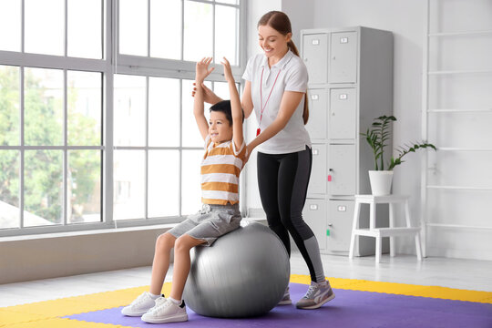 Cute little Asian boy training on fitball with coach in gym