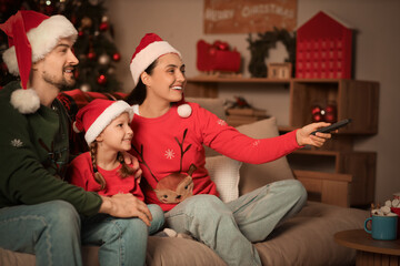 Little girl with her parents in Santa hats watching Christmas movie on sofa at home