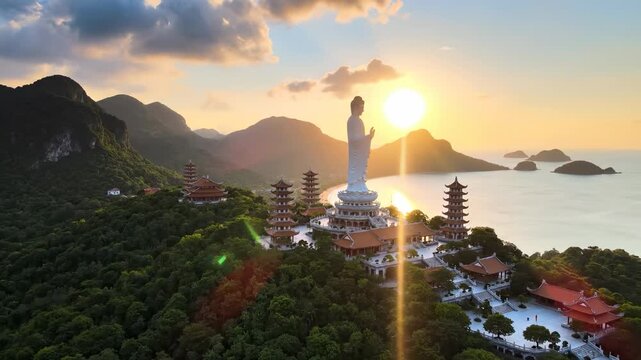 A tall statue of a serene figure overlooks the mountains and ocean at sunset, creating a tranquil atmosphere. The statue and surrounding temples are embraced by lush greenery and h