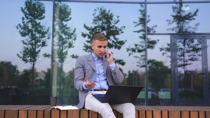 Fototapeta premium Angry man working with laptop, talking on the phone, sit on bench. Slow motion