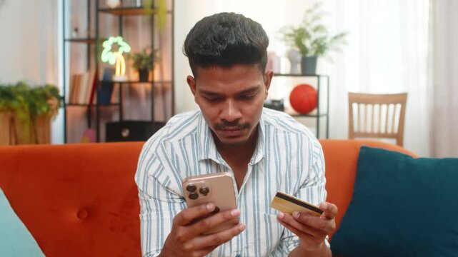 Indian man at home uses smartphone plus credit card to place order enters cvv and smiles after checkout. Arabian guy on sofa confirms order sees app status waits for delivery and feels safe payment.