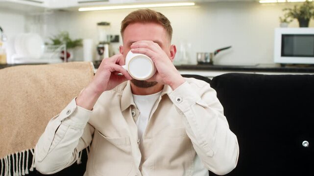Young man at home holds cup of tea warms hands smiles and breathes herb aroma during a short pause. Caucasian guy on sofa enjoys calm break sips tea and shows peace comfort warm and relaxed soft mood.