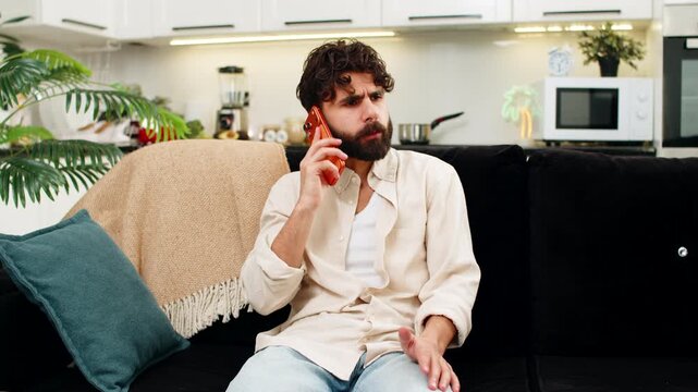 Indian man at home looks at smartphone gets spam call feels annoyed frowns ends call quickly. Arabian guy on sofa leans on pillow holds mobile phone to chest feels tired anger after loud ring inside.