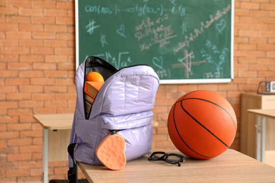 Stylish school backpack with stationery, ball and eyeglasses on desk in classroom