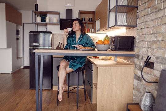 Beautiful young woman in silk robe having morning coffee with smile face, enjoying, closed eyes, sitting at the kitchen. Morning routine, domestic life