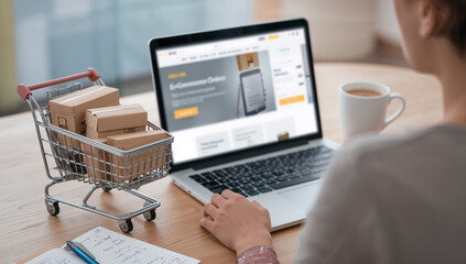 Person shopping online with a shopping cart and cardboard packages next to a laptop