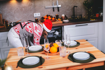 Woman unloading dishwasher in Christmas and New Year kitchen with fully prepared holiday table, warm lights, decor and pinecones creating cozy winter atmosphere for seasonal festive celebration.