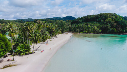 Serene beach on Koh Kood Island in Thailand showcases soft sands, swaying palm trees, and turquoise waters. Visitors relax along the shore, enjoying the tropical paradise under a clear sky.