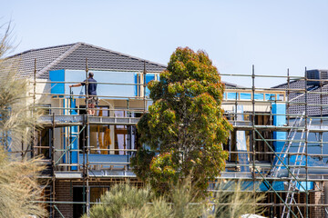 A residential construction site in a suburban neighborhood in Australia, showing a modern house under construction with scaffolding, protective building wrap, and a worker on site. New housing growth © Doublelee