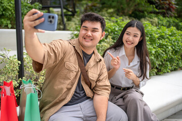 Smiling Asian friends taking a smartphone self-portrait. They hold retail bags outside a contemporary high-rise after a successful shopping trip.